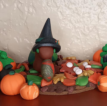 Polymer clay sculpture of a witch snail among leaves and pumpkins. White runes cast over leaves. Black caterpillar familiar on her hat.