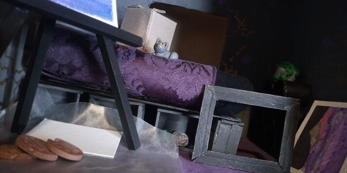 Photo of the inside of a miniature room box styled as a goth bedroom with black & purple. Painter's tools strewn across floor and a small grey cat resting on the bed next to a box. A green plant barely visible in the background.